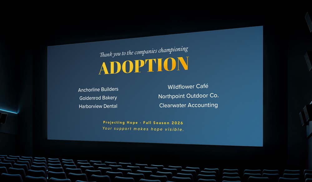 A cinema screen viewed from the back of the theater over rows of seats — your name + category appear up there.